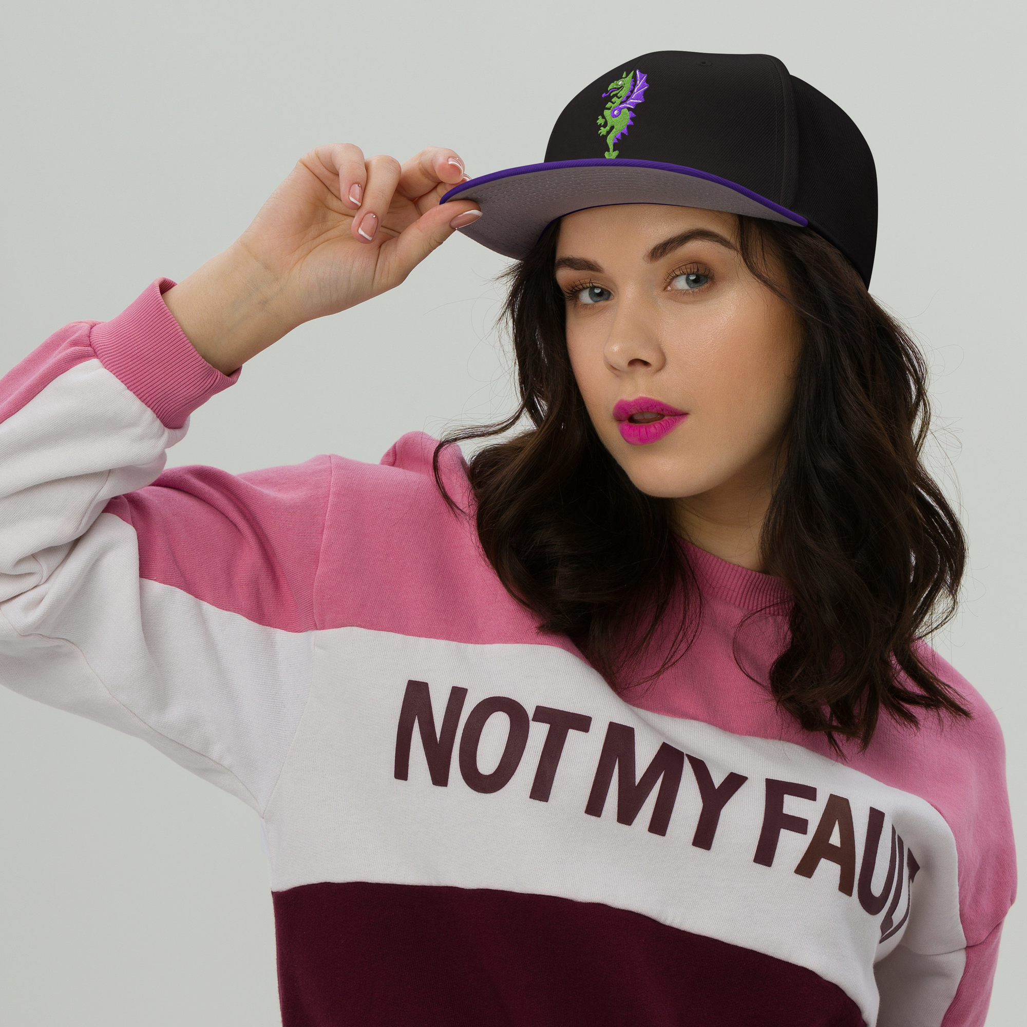 Fashionable female model wearing a black flat bill hat with a bright purple bill and strap featuring embroidered dragon master remix design with matching bright purple wings. Item is a BRKDrip AFOL Headgear Dad Hat.