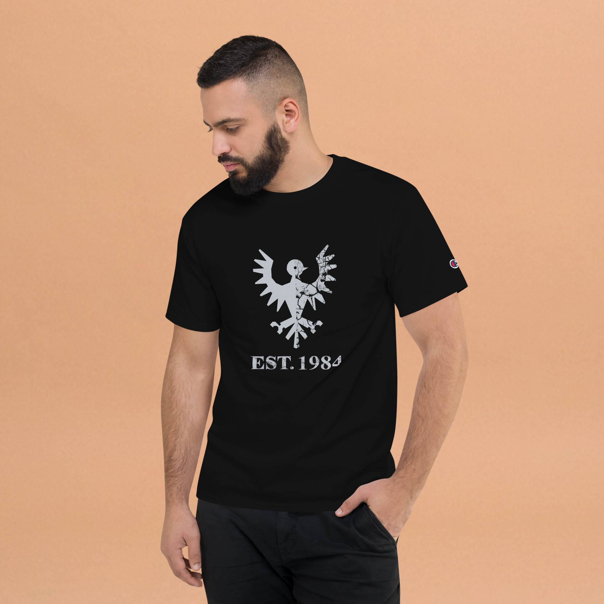 Fashionable male model against orange background wearing black heavyweight, 100% cotton luxury Champion© t-shirt featuring aged falcon emblem and text "EST. 1984" indicating the year the faction was first introduced to the brick universe. Item is a BRKDrip Heritage Collection luxury shirt with a vintage castle building block aesthetic.