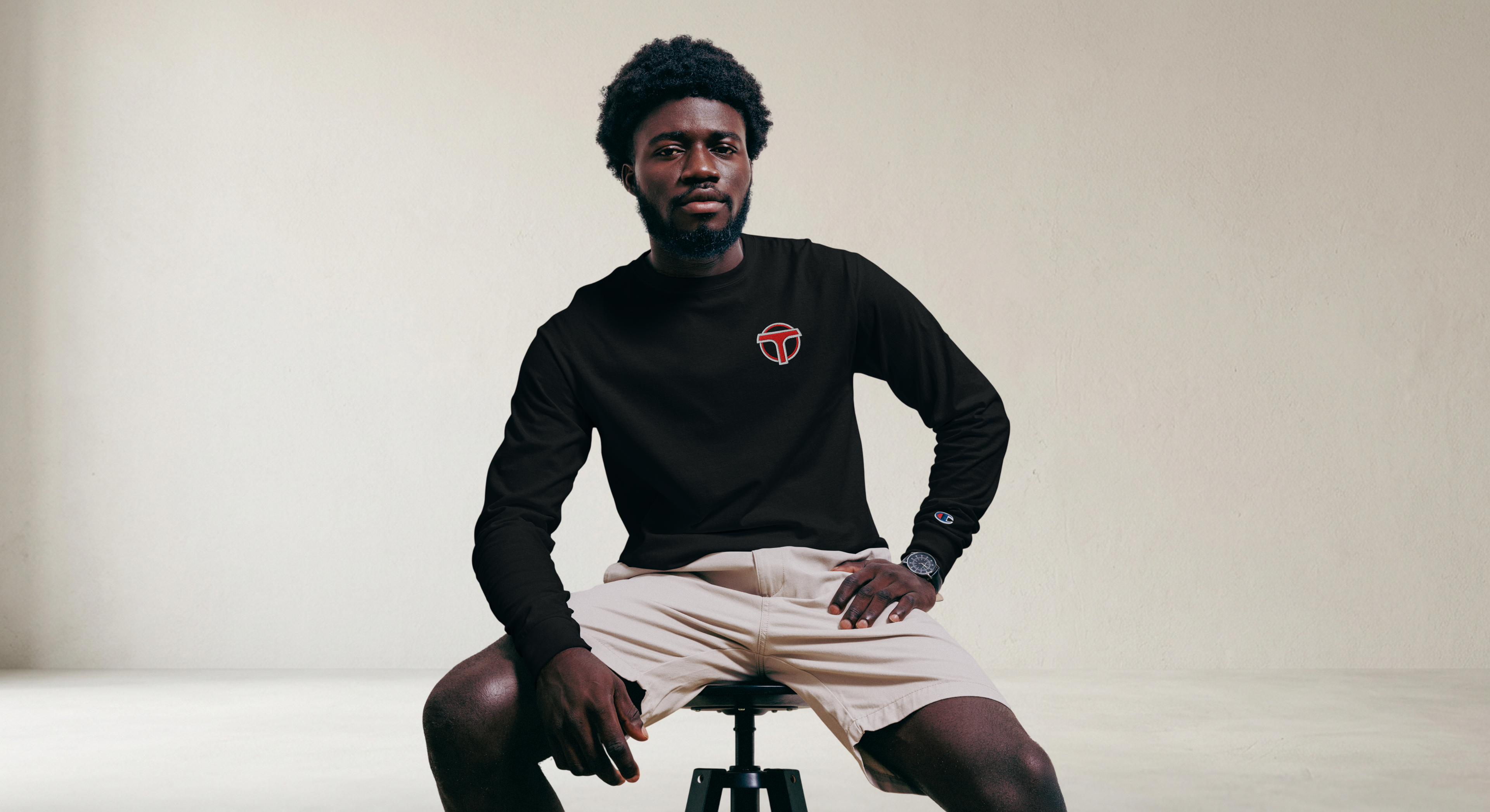 Fashionable male model sitting on a stool wearing a black lightweight, 100% cotton luxury Champion© shirt featuring airport shuttle emblem embroidered in the top right corner. Item is a BRKDrip Builder's Collection luxury longsleeve shirt with a classic town building block aesthetic.