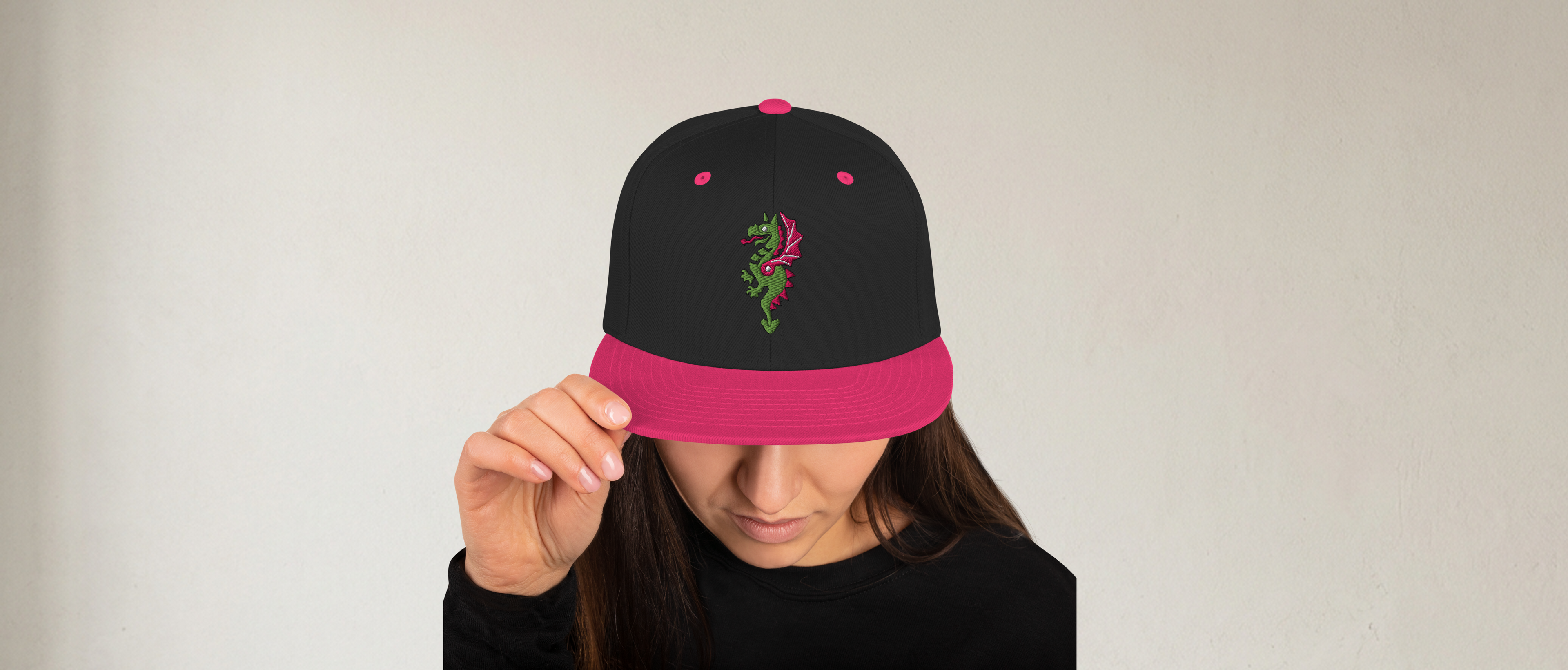 Fashionable female model wearing a black flat bill hat with a bright pink bill and strap featuring embroidered dragon master remix design with matching bright purple wings. Item is a BRKDrip AFOL Headgear classic snapback with a vintage castle building blocks aesthetic.
