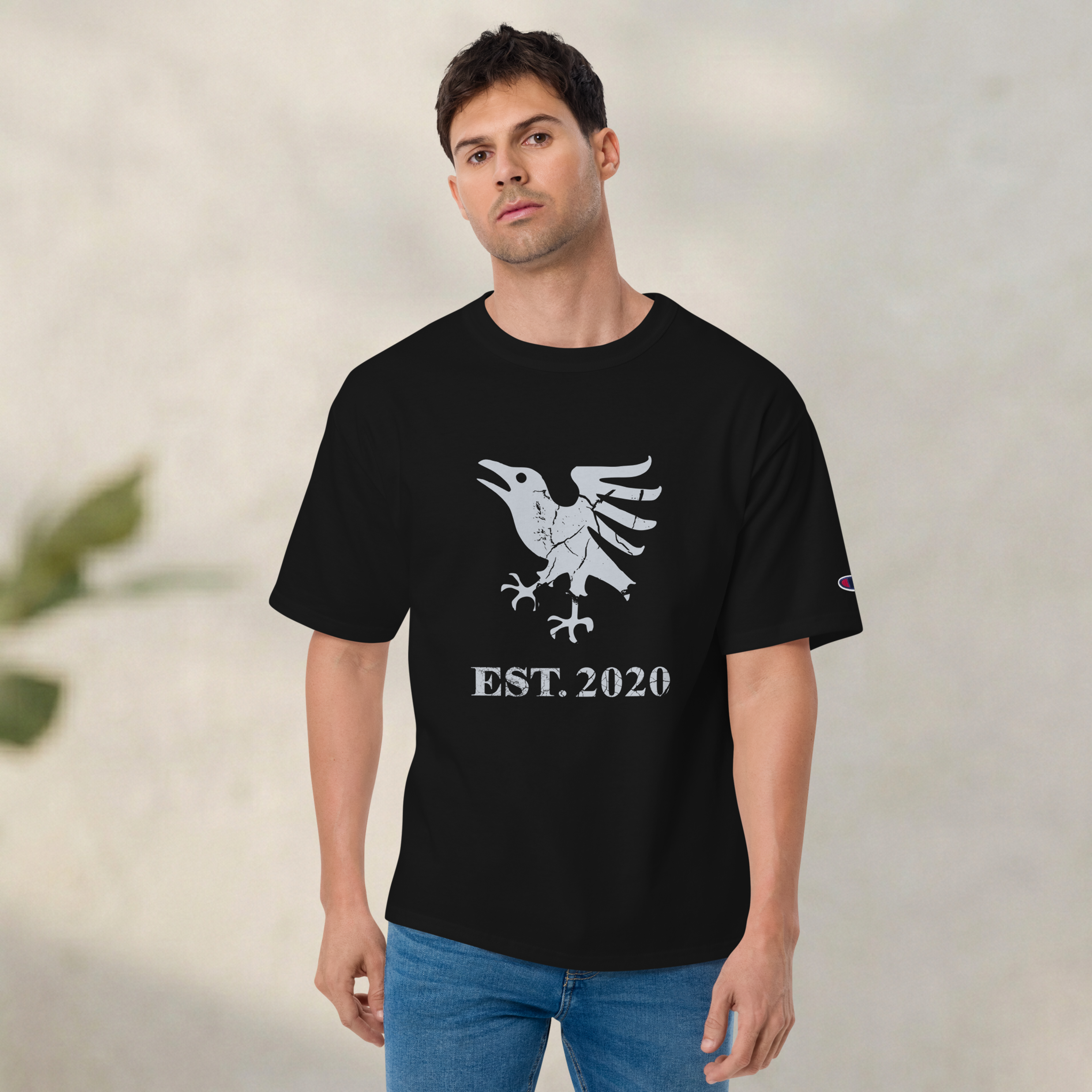 Fashionable male model on grey background wearing black heavyweight, 100% cotton luxury Champion© t-shirt featuring aged raven emblem and text "EST. 2020" indicating the year the faction was first introduced to the brick universe. Item is a BRKDrip Heritage Collection luxury shirt with a vintage castle building block aesthetic.